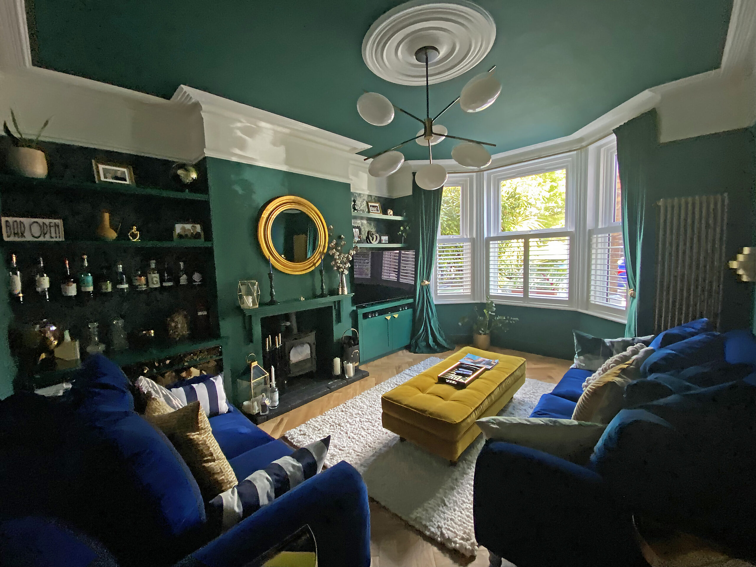 Colour-drenched living room in deep green with navy velvet sofas, a mustard velvet ottoman, white cornicing and a bay window with shutters