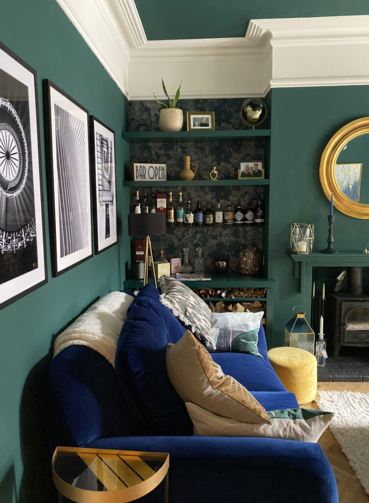 Built-in alcove shelves painted dark green with a dark patterned wallpaper behind, a navy velvet sofa, a round gilt mirror and black-and-white framed prints