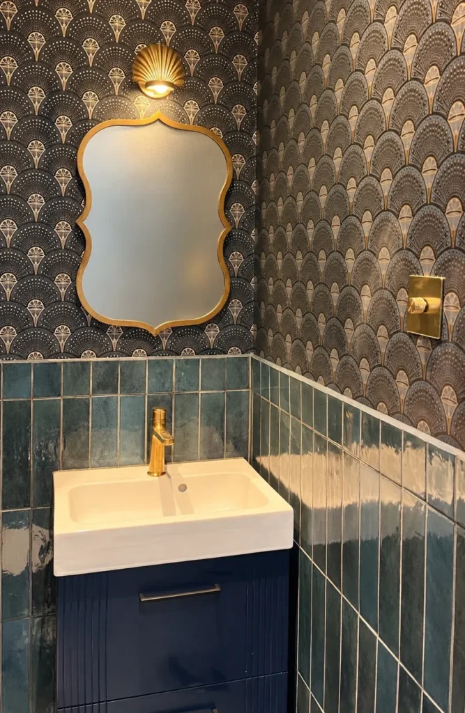 Small downstairs toilet with navy and gold Art Deco fan wallpaper, glossy teal vertical tiles, a navy vanity unit, a brass tap and a scalloped gold mirror under a brass shell wall light