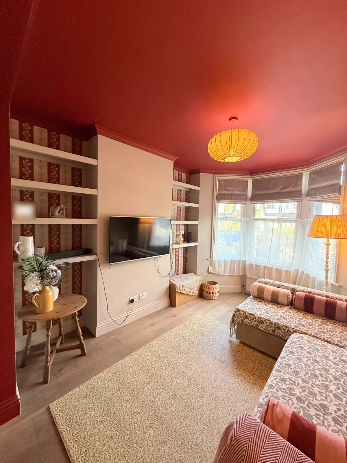 The finished front room in a North London Victorian terrace: a red ceiling, floral striped wallpaper behind the alcove shelves, a pleated pendant light, a bay window with blinds and voiles, a patterned rug and the existing sofa dressed with floral throws and striped bolster cushions