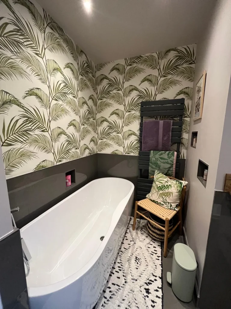 Freestanding bath surrounded by green palm leaf wallpaper above dark grey tiles, with a charcoal towel radiator, a woven chair, coordinating towels and framed art
