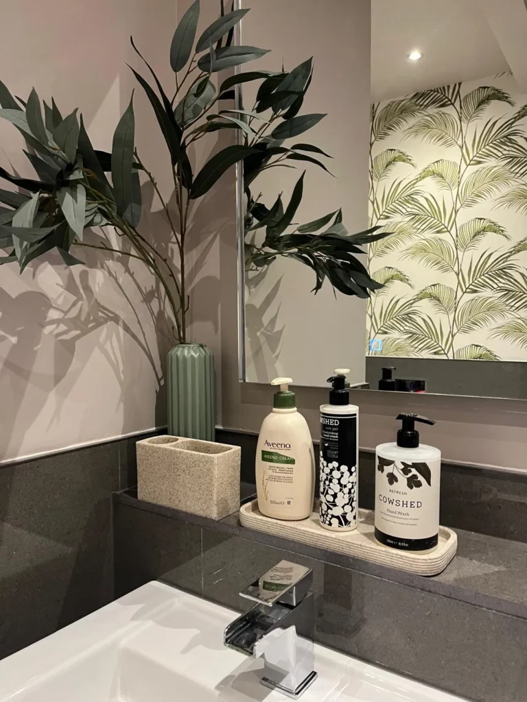 Soft mauve bathroom wall with eucalyptus stems in a sage green vase, a stone-effect tray and toothbrush holder, and palm leaf wallpaper reflected in the mirror