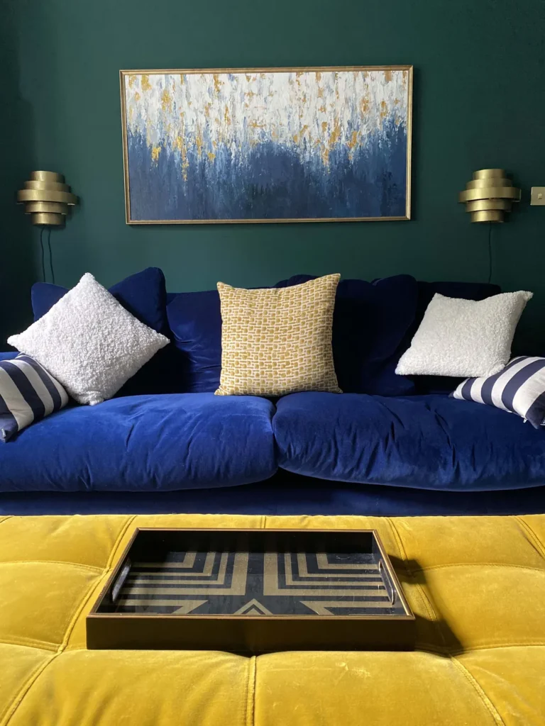Navy velvet sofa with bouclé and striped cushions under an abstract blue and gold painting, flanked by tiered brass Art Deco wall lights, with a mustard velvet ottoman and geometric tray