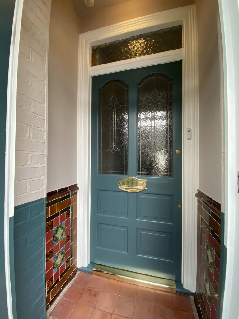 Inner front door painted deep teal with leaded glass panels and a polished brass letterbox, framed by original patterned tiles, teal-painted brickwork and a quarry tile floor